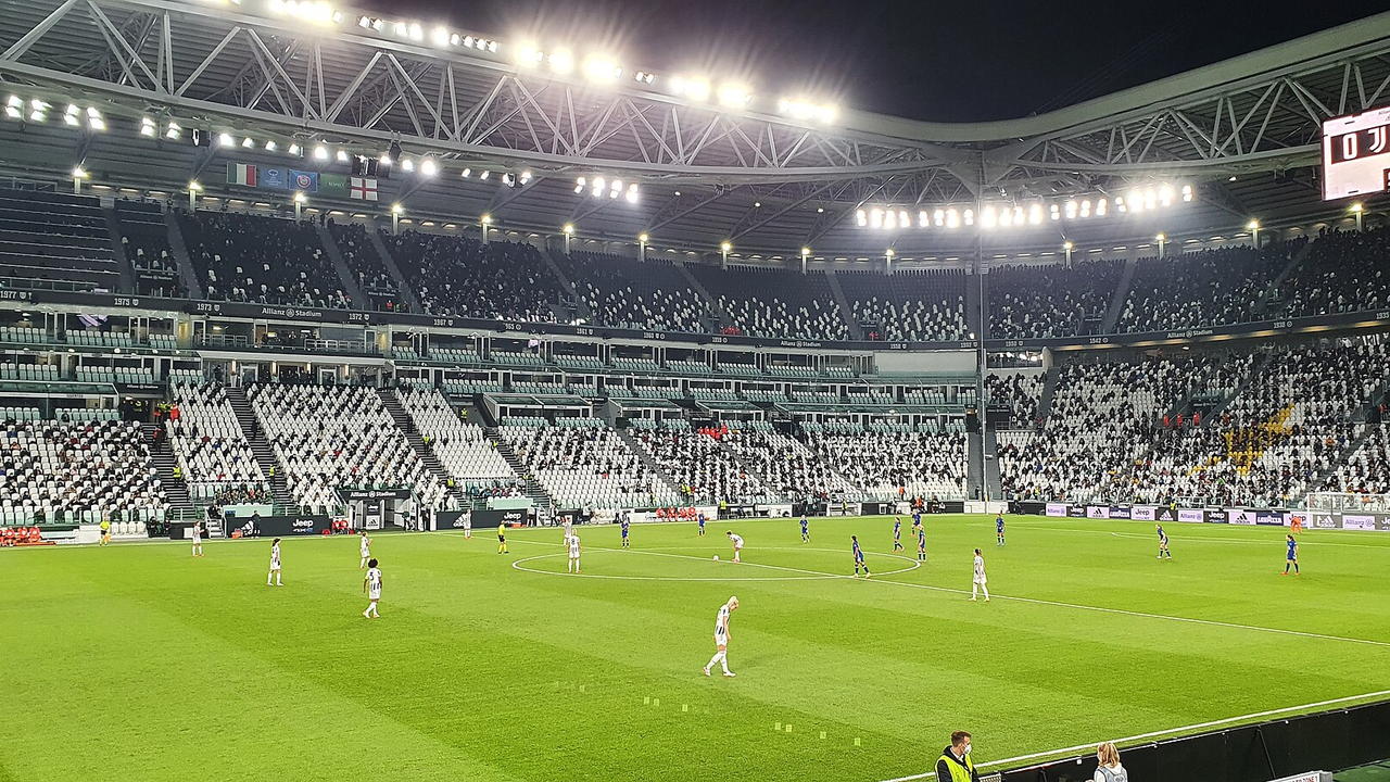 Allianz Stadium in Turin — Blick von der Tribüne über den Rasen, Flutlicht an, die Ränge in Schwarz und Weiss gehalten