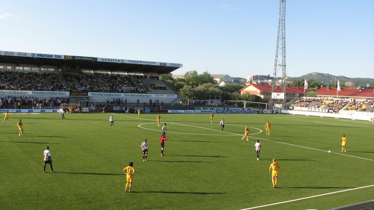 Aspmyra Stadion in Bodø — Spielszene bei Tageslicht, niedrige überdachte Tribüne und ein Flutlichtmast, dahinter Hügel