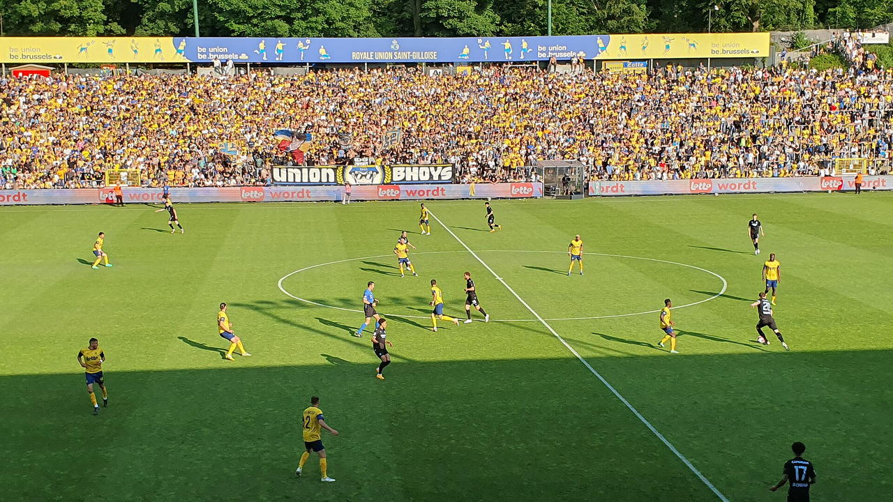 Stade Joseph Mariën in Brüssel — Spielszene bei Tageslicht, dicht besetzte Ränge in Gelb und Blau, dahinter Baumbestand